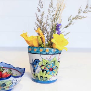 May include: A hand-painted ceramic vase with a blue rim and floral design, holding yellow lilies, purple flowers, and dried grasses. A matching bowl with strawberries is in the foreground.
