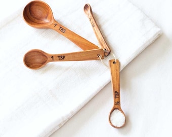 May include: A set of three wooden measuring spoons with a white cloth background. The spoons are labelled "1 TBS", "1 TSP", and "1/2 TSP".