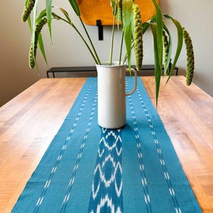 May include: A white ceramic pitcher with a handle, filled with green foliage and seed pods, sits on a blue and white patterned table runner.