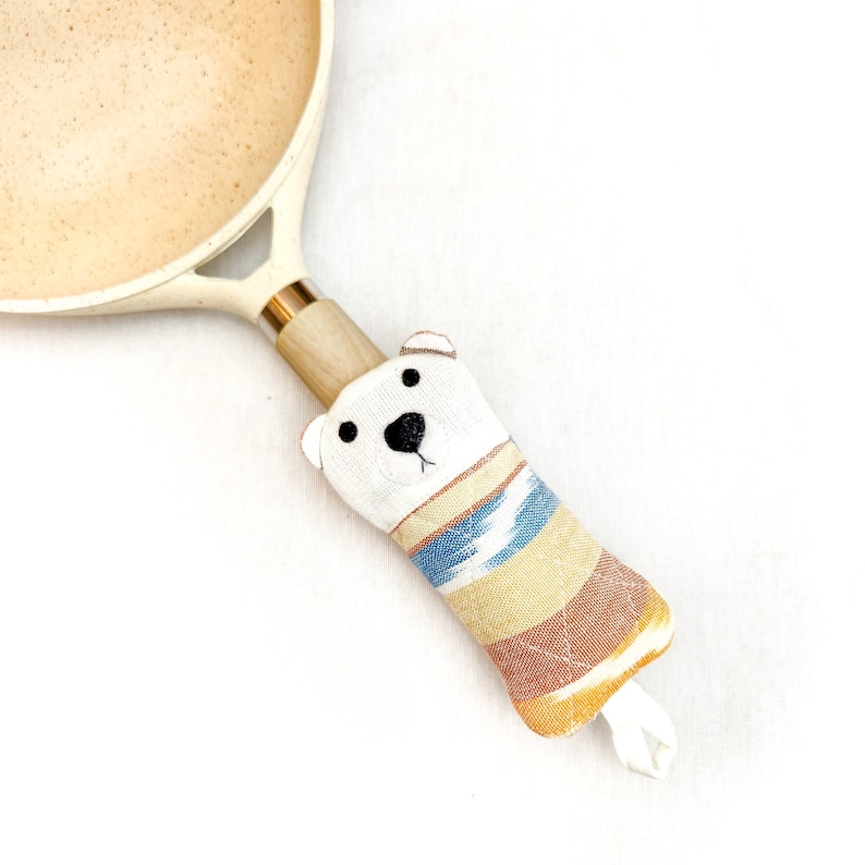 May include: A white fabric pot holder with a bear face and a striped pattern in shades of blue, yellow, and orange. The pot holder is attached to a wooden handle.