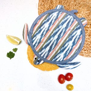 May include: A blue and white striped fabric turtle pot holder with a brown and pink pattern. The pot holder is on a woven straw mat.
