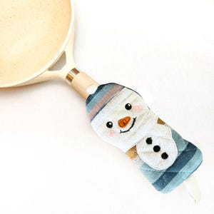 May include: A cream-colored frying pan with a light wood-tone handle covered by a fabric handle sleeve. The sleeve features a snowman design with a blue and white striped hat, an orange carrot nose, black eyes, and three black buttons.