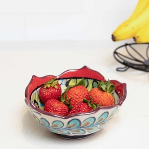 May include: A red and white ceramic bowl with a floral design and a scalloped rim. The bowl is filled with fresh strawberries.