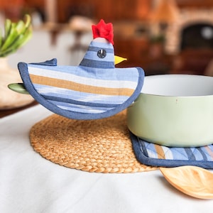 Könnte beinhalten: Ein blauer und gelber gestreifter Topflappen in Form eines Huhns mit rotem Kamm. Der Topflappen liegt auf einer geflochtenen Strohmatte mit einem grünen Keramiktopf daneben.