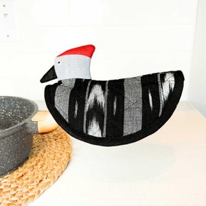 May include: A whimsical bird-shaped pot holder with a red, gray, and black color scheme. The bird's head is gray with a red crest and black beak. The body features a black and white patterned design. This kitchen accessory is designed to protect hands from hot cookware.