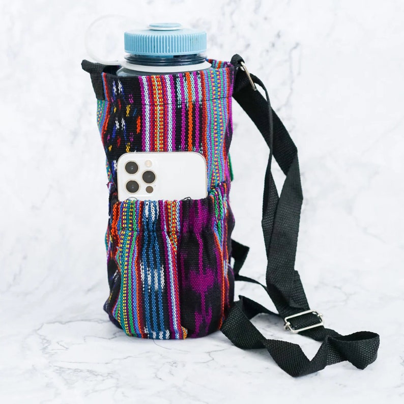 May include: A black and multi-colored fabric water bottle holder with a black adjustable strap. The holder has a pocket for a phone and a zippered pocket. A clear water bottle is inside the holder.