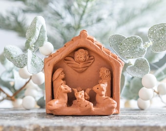 Puede incluir: Un belén de terracota con un pequeño establo de arcilla, María, José y el niño Jesús. Las figuras están rodeadas por dos animales de arcilla.
