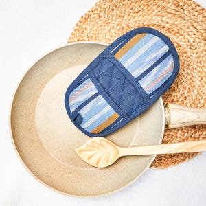 May include: A blue and white striped pot holder with a quilted denim center. The pot holder is on top of a brown woven mat and a light brown frying pan.