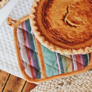 Puede incluir: Una tarta de calabaza casera con una corteza dorada se encuentra sobre una almohadilla acolchada de rayas de colores.