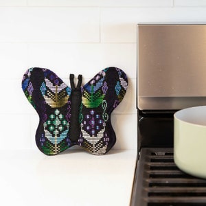 May include: A black butterfly-shaped oven mitt with colorful embroidered designs in shades of green, blue, purple, and gold. The mitt is sitting on a white countertop next to a black and silver stove.
