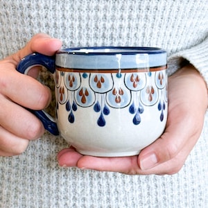 Puede incluir: Una taza de cerámica con asa y borde azules, con un cuerpo blanco adornado con un patrón repetido de formas de gotas azules y marrones. La taza está sostenida en las manos de una persona.