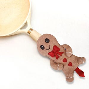 May include: A brown gingerbread man pot handle cover with a red bow and heart accents. The cover is attached to a light-colored pan with a wooden handle. The gingerbread man has black eyes and a smiling face. A festive kitchen accessory.