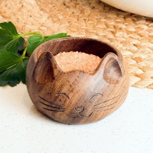 May include: A wooden salt cellar shaped like a cat with a smiling face. The cat is holding pink Himalayan salt inside.