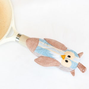 Puede incluir: Un agarrador de olla con forma de búho azul y marrón con un patrón de rayas blancas y amarillas. El agarrador de olla está unido a un mango de sartén blanco y plateado.