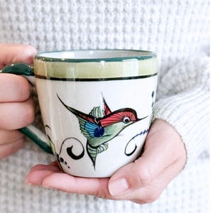 May include: A ceramic mug with a hand-painted hummingbird design. The mug has a white base, a green rim and handle. The hummingbird is depicted with red, blue, and green details, and is surrounded by black swirls.