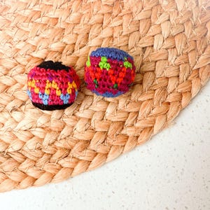 May include: Two small, spherical, crocheted items featuring intricate yarn work. The left item displays a pattern of black, red, yellow, and light blue. The right item features a vibrant design of purple, red, and green yarn in a repeating motif.