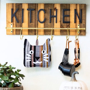 May include: A wooden sign with the word "KITCHEN" in black letters hangs on a wall with a gold metal rack with hooks. A cat-shaped pot holder, measuring cups, and a copper whisk hang from the hooks.