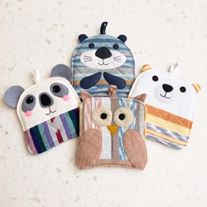May include: Four quilted fabric pot holders shaped like animals. One is a gray and white koala, one is a blue striped otter, one is a brown and tan owl, and one is a white bear with yellow and orange stripes. Each has a small hanging loop.