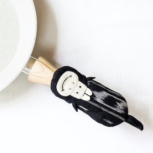 May include: Black and white patterned fabric pot holder with a smiling white face and black eyes. The pot holder is attached to a wooden handle.