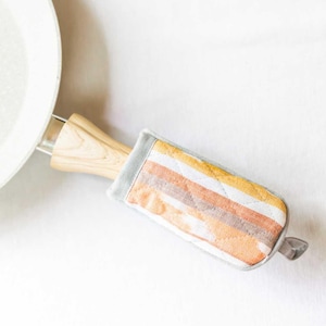 May include: A striped fabric pot holder with a light gray background and orange, yellow, and brown stripes. The pot holder is wrapped around the handle of a white frying pan.