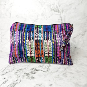 Puede incluir: Una bolsa con cremallera colorida con un patrón tejido en tonos de morado, azul, verde, naranja y blanco. La bolsa tiene un tirador de cremallera con borla.