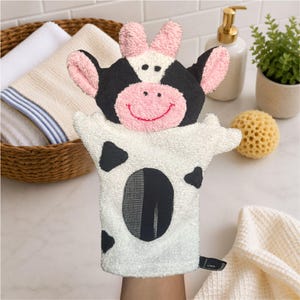 May include: A cow-shaped bath mitt made of white, black, and pink textured terry cloth fabric. The mitt features a cow face with pink ears, a pink snout, and black patches on the body. It is designed to be worn on the hand for washing.