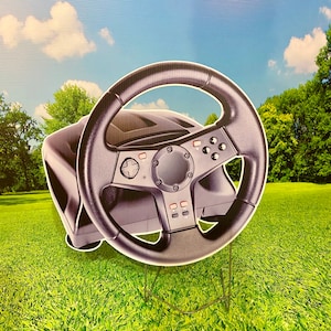 May include: A black plastic racing wheel with buttons and a steering wheel. The wheel is on a stand and is in front of a green grassy background.