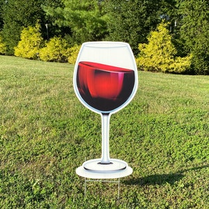 Puede incluir: Un gran recorte de cartón blanco de una copa de vino llena de vino tinto. La copa está de pie sobre un césped verde.