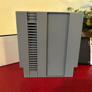 May include: A gray plastic cartridge case for a Nintendo Entertainment System (NES) video game console. The case has a slot for the game cartridge and a label on the front.