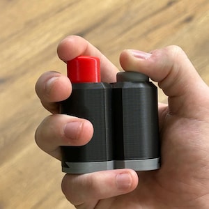 May include: A black, rectangular device with two cylindrical buttons, one red and one gray. The device is held in a hand, with the buttons positioned for pressing. The base of the device is gray.