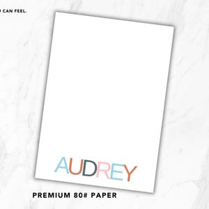 May include: A rectangular sheet of white 80# paper featuring the name AUDREY printed in multicolored sans-serif letters at the bottom. The text QUALITY YOU CAN FEEL. SMALL DETAIL. BIG DIFFERENCE. and PREMIUM 80# PAPER are printed on the page.