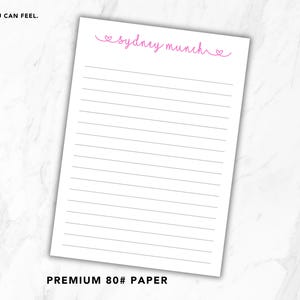 May include: A white notepad sheet featuring horizontal black lines and the pink cursive text "sydney munch" flanked by two small heart icons at the top. The paper is labelled as premium 80lb paper with the text "QUALITY YOU CAN FEEL. SMALL DETAIL. BIG DIFFERENCE."