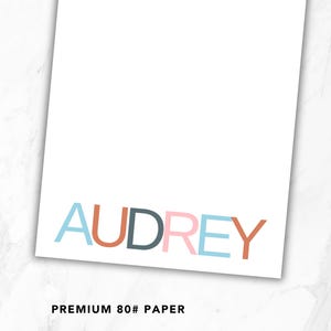 May include: A rectangular sheet of white 80# paper featuring the name AUDREY printed in large, sans-serif capital letters. Each letter is colored differently in shades of light blue, terracotta, dark gray, and soft pink, arranged horizontally.