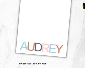May include: A white sheet of premium 80# paper featuring the name AUDREY printed in large, sans-serif capital letters. Each letter is a different colour, including light blue, terracotta, dark grey, and soft pink, arranged horizontally.