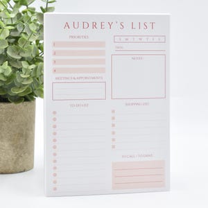 May include: A white paper notepad titled Audrey's List in red text, featuring sections for priorities, meetings, appointments, notes, a to-do list with circular bullets, a shopping list with square checkboxes, and a to-call or to-email section.