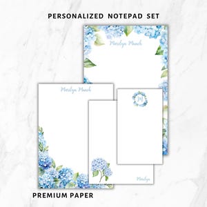 May include: A personalized notepad set featuring a floral design with blue hydrangeas and green leaves. The set includes various sizes of notepads with the text "Marilyn Munch" and "Premium Paper".