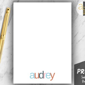 May include: A white stationery sheet with the name "audrey" in colorful letters at the bottom. A gold pen rests on the marble surface next to the sheet. A black and white logo is in the upper right corner, and a "PROOF" stamp is in the lower right corner.