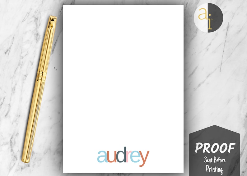 May include: A white stationery sheet with the name "audrey" in colorful letters at the bottom. A gold pen rests on the marble surface next to the sheet. A black and white logo is in the upper right corner, and a "PROOF" stamp is in the lower right corner.