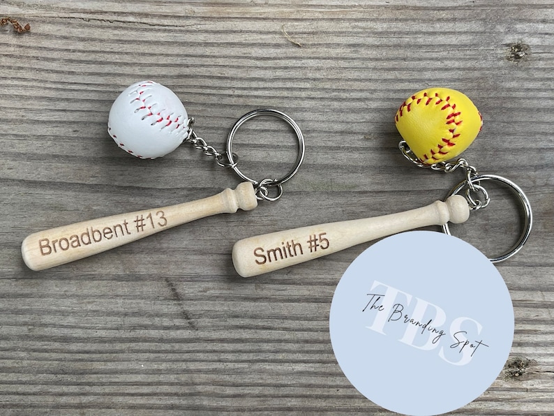 May include: Two personalized wooden baseball bat keychains with a white baseball and a yellow softball. The bat keychains are engraved with the names "Broadbent #13" and "Smith #5". The keychains are attached to a white circle with the text "The Branding Spot" in black.