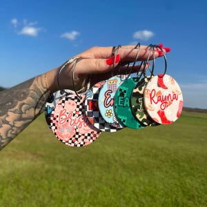 May include: A hand holds six round, personalized luggage tags with silver metal loops. The tags feature various designs, including a pink and black checkered pattern, a Minecraft-themed design, and a cowgirl boot design with the name "Rayna".