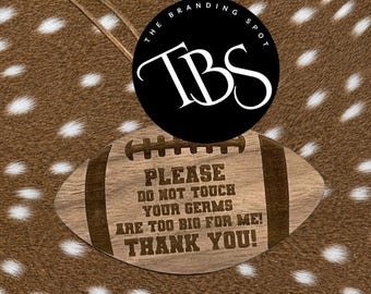 May include: A wooden football-shaped sign with the text "PLEASE DO NOT TOUCH YOUR GERMS ARE TOO BIG FOR ME! THANK YOU!" and a string for hanging. The sign has a light brown color and a dark brown border. The Branding Spot logo is in the background.