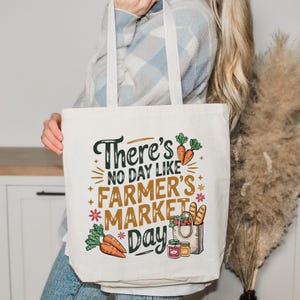 May include: Cream-colored tote bag with the text "There's no day like Farmer's Market Day" in green, orange, and brown. The bag features illustrations of carrots, flowers, bread, and jars of jam, perfect for carrying groceries.