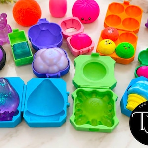 May include: A collection of colorful silicone molds and squishy toys in various shapes including a purple bear, a bright orange sphere, a pink dumpling, and a blue ice cream cone. Several open plastic cases reveal molded shapes in vibrant colors.
