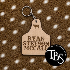 May include: A wooden keychain with a cow tag design. The tag features the name "RYAN STETSON MCCALL" engraved in a bold font, with a cow silhouette above. A metal keyring and chain are attached. The Branding Spot logo is in the lower right.