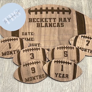 May include: Wooden football-shaped milestone marker set. The set includes a large football with engraved text and smaller footballs marked 1, 3, 5, 7, 9 months, and 1 year. The large football has space for date, time, and weight.