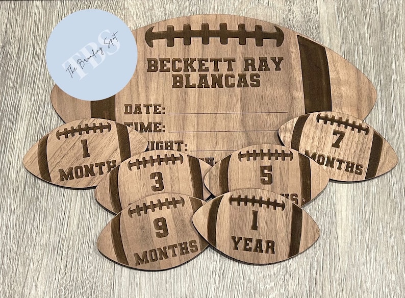 May include: Wooden football-shaped milestone marker set. The set includes a large football with engraved text and smaller footballs marked 1, 3, 5, 7, 9 months, and 1 year. The large football has space for date, time, and weight.