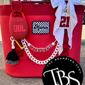 May include: A red cooler bag with a JBL speaker, a "Mamas" patch, and a white chain with the name "Makayla #21". A white bow and a tag with the number "21" are attached. The Branding Spot logo is visible.