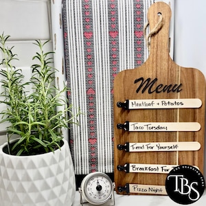 May include: Wooden menu board with the word "Menu" in black script. The board has four slots holding menu options: "Meatloaf + Potatoes", "Taco Tuesday", "Fend For Yourself", "Breakfast For", and "Pizza Night". A timer and a potted plant are in the background.