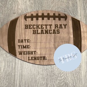 May include: A wooden football-shaped sign with engraved details. The sign includes the name "BECKETT RAY BLANCAS" and spaces for date, time, weight, and length. The sign is light brown with darker brown accents.