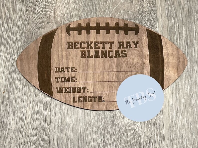 May include: A wooden football-shaped sign with engraved details. The sign includes the name "BECKETT RAY BLANCAS" and spaces for date, time, weight, and length. The sign is light brown with darker brown accents.
